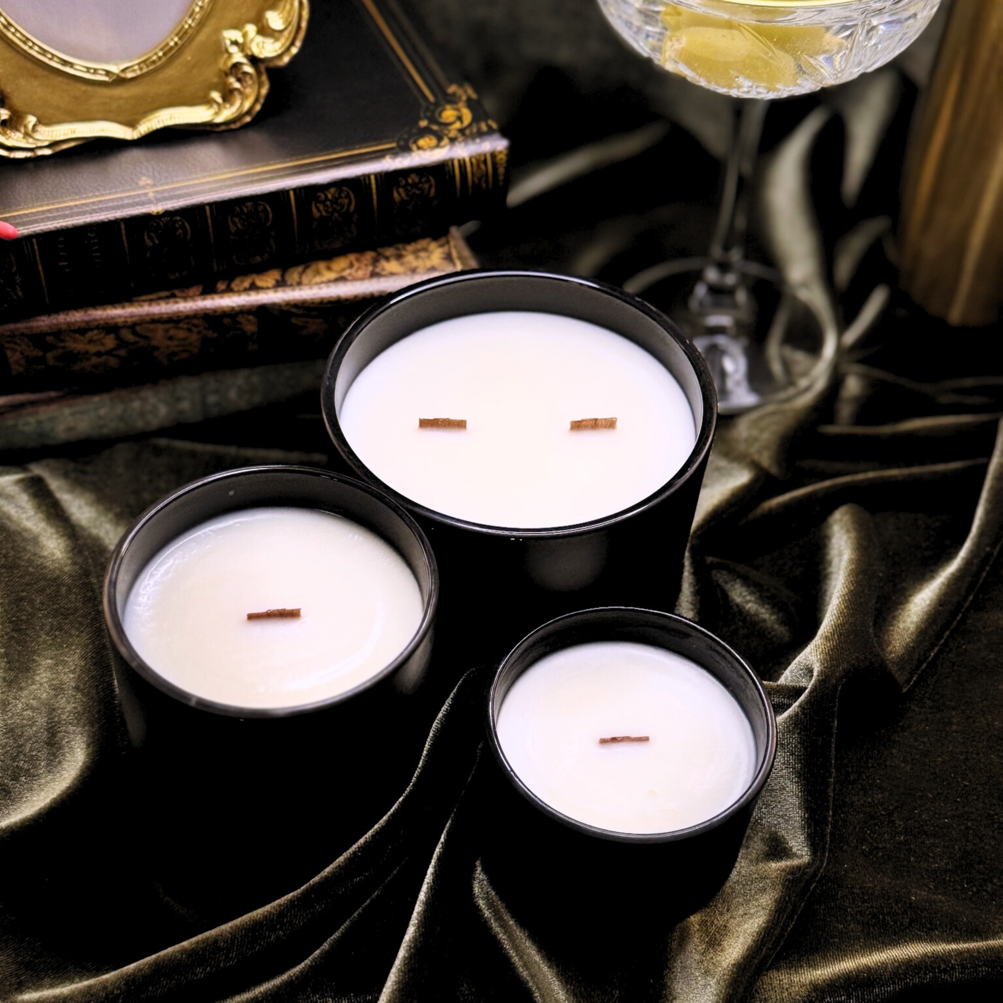Three black candles with white wax on a dark fabric surface, with a glass of white wine and decorative books in the background.
