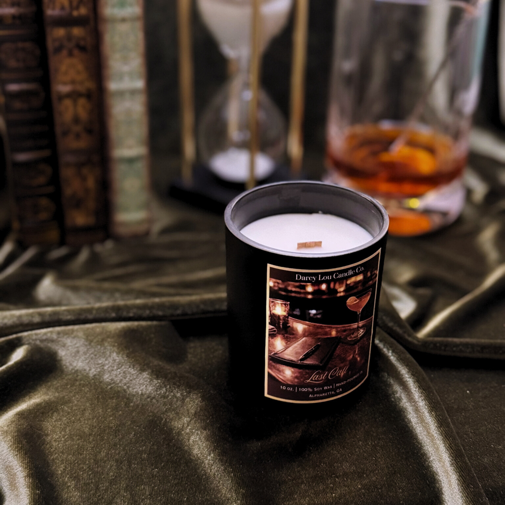 Candle in a black tin with a label on a dark surface, with glasses and books in the background.