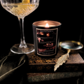 Candle with a decorative label next to a glass of white wine on a dark surface